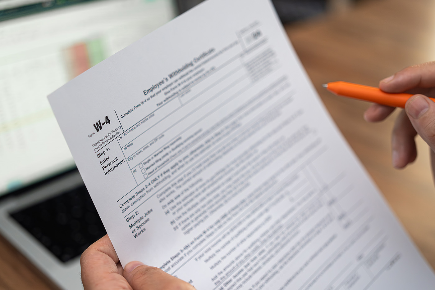 Newly hired employee compares data in modern laptop to fill W-4 form to determine federal tax withholding for new job sitting at desk in office with pen.